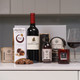 Pamper Night In Treat Box. A selection of gourmet items including a bottle of red wine labeled "Montlort" a box of sticky toffees, a hand scrub, and scented candles. A glass of red wine sits beside the products on a white surface. The background is softly blurred, focusing attention on the arranged items. Pamper Night In Treat Box. A selection of gourmet items including a bottle of red wine labeled "Montlort" a box of sticky toffees, a hand scrub, and scented candles. A glass of red wine sits beside the products on a white surface. The background is softly blurred, focusing attention on the arranged items.